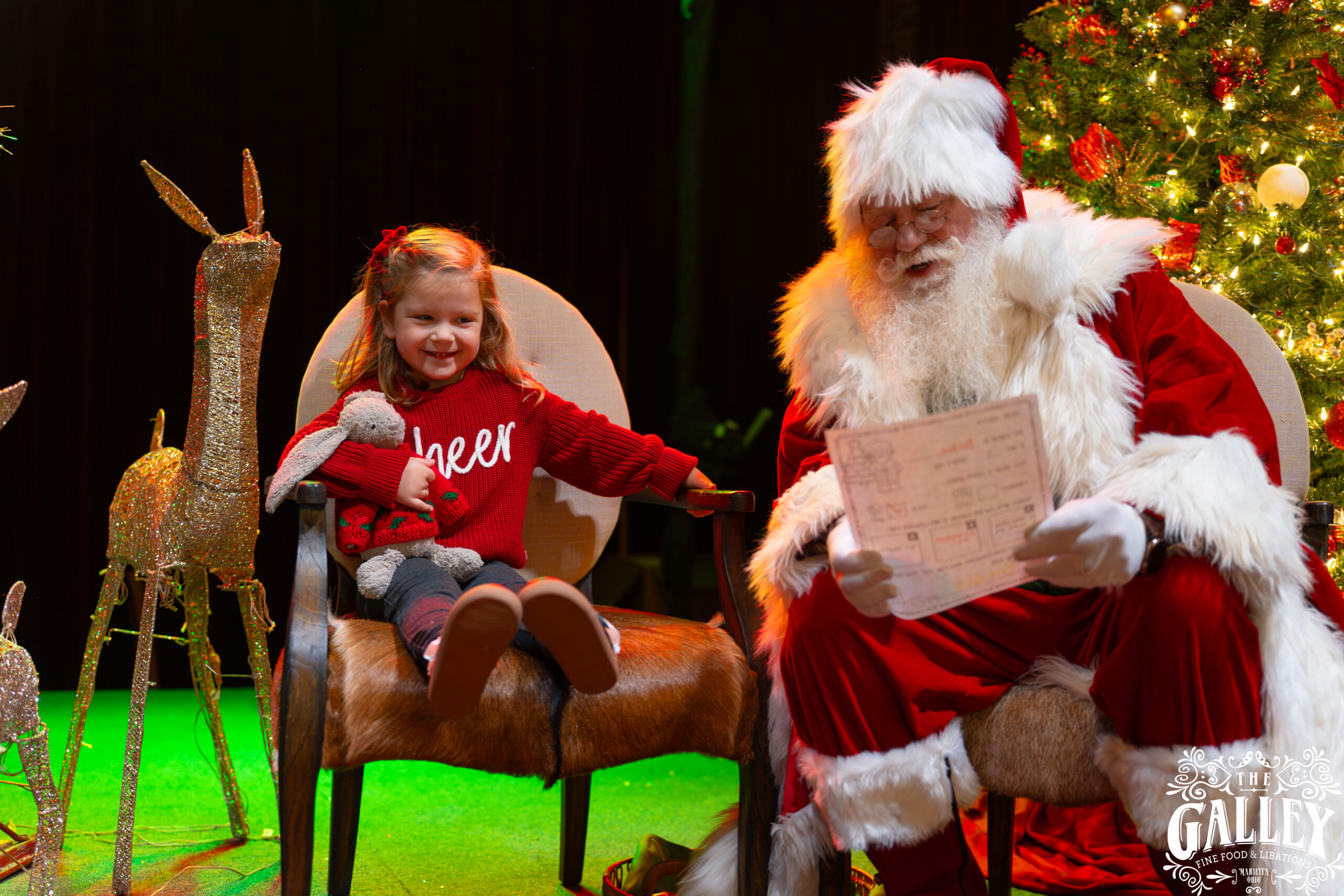 A child holding a stuffed rabbit sits beside Santa at The Galley